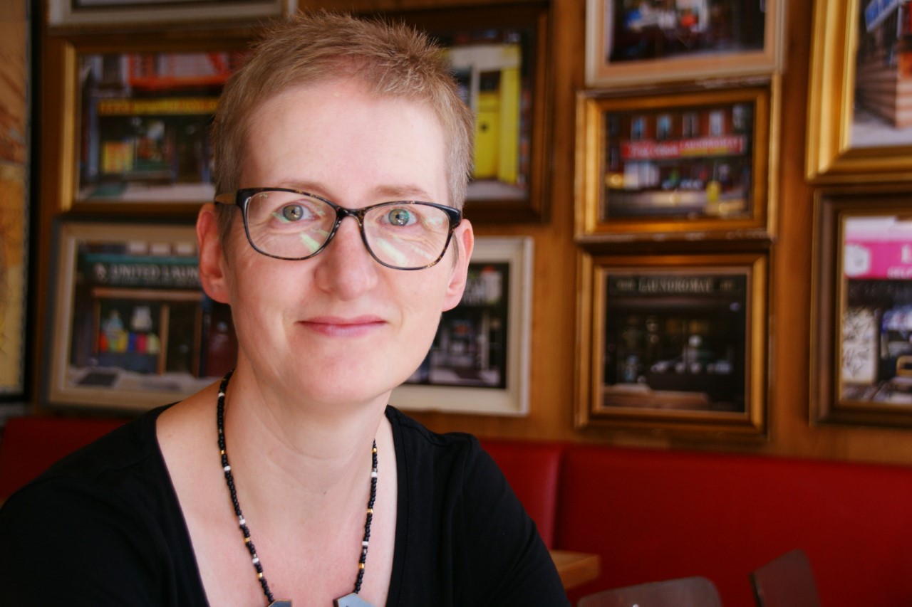 Dr Williams sitting in a room with painting covering the wall, wearing a black top, necklace and glasses.