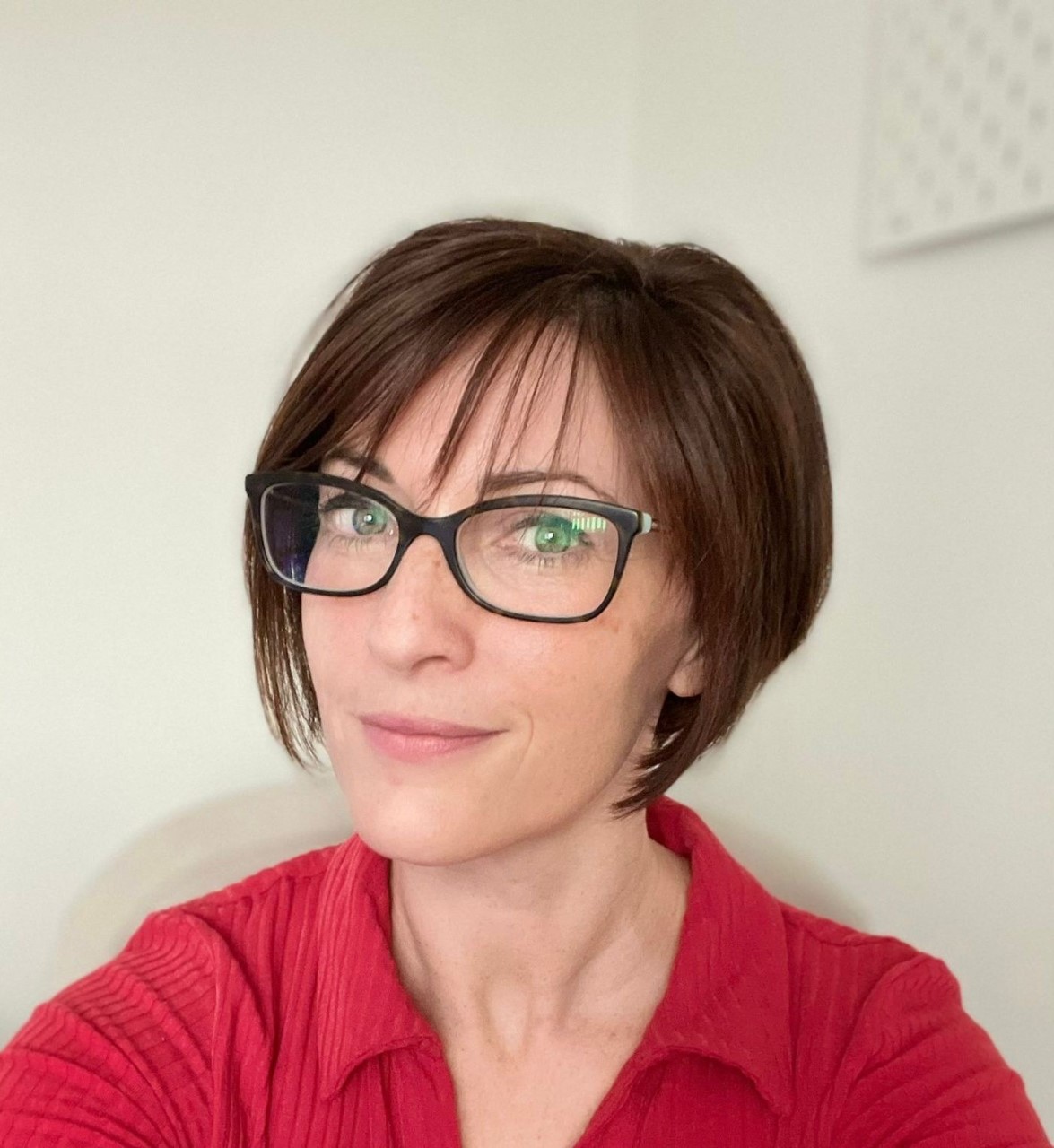 Dr Packard in front of a white wall wearing a red top and black glasses